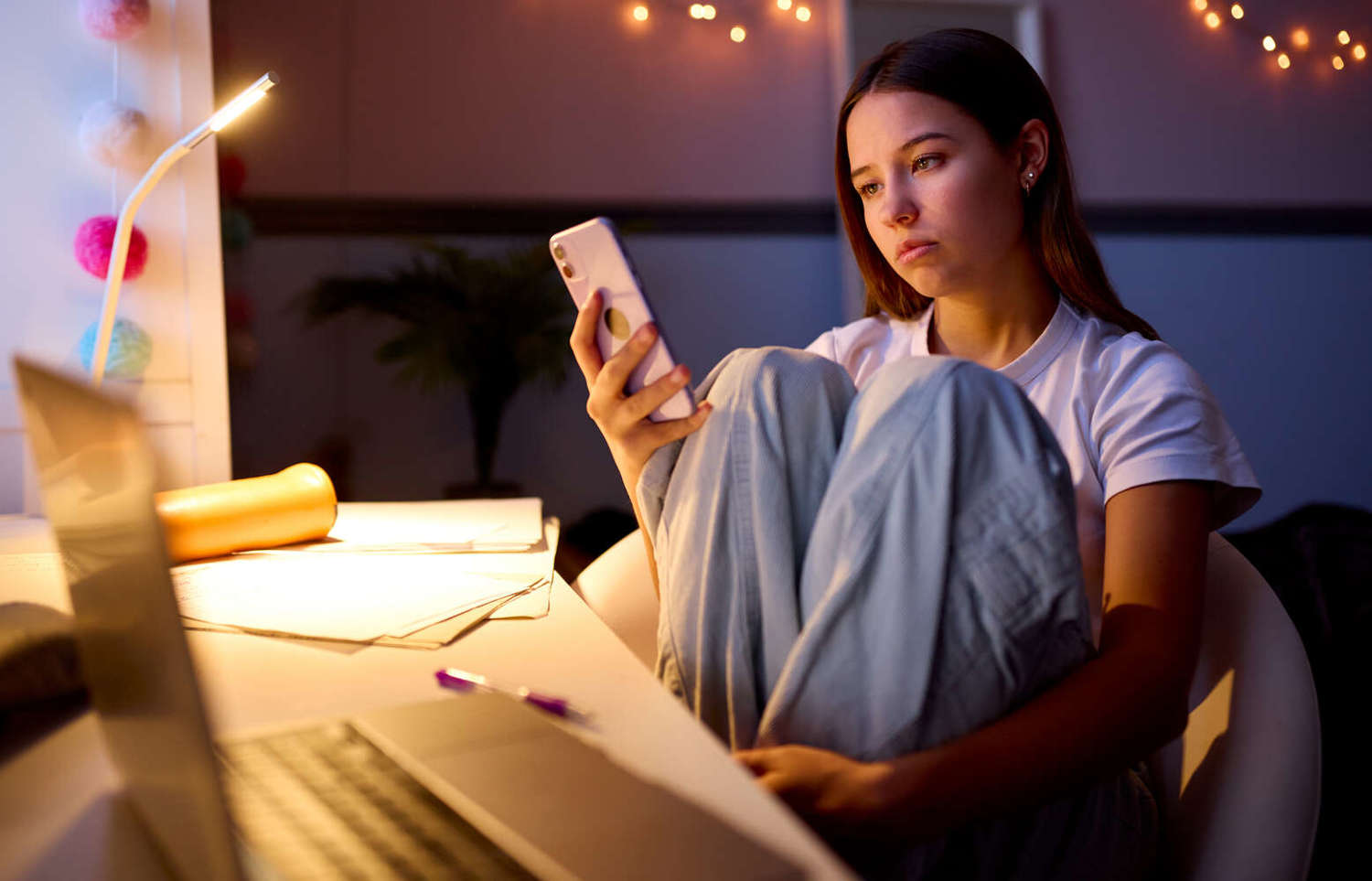 Worried Teenage Girl Sitting At Desk In Bedroom At Home Looking At Mobile Phone